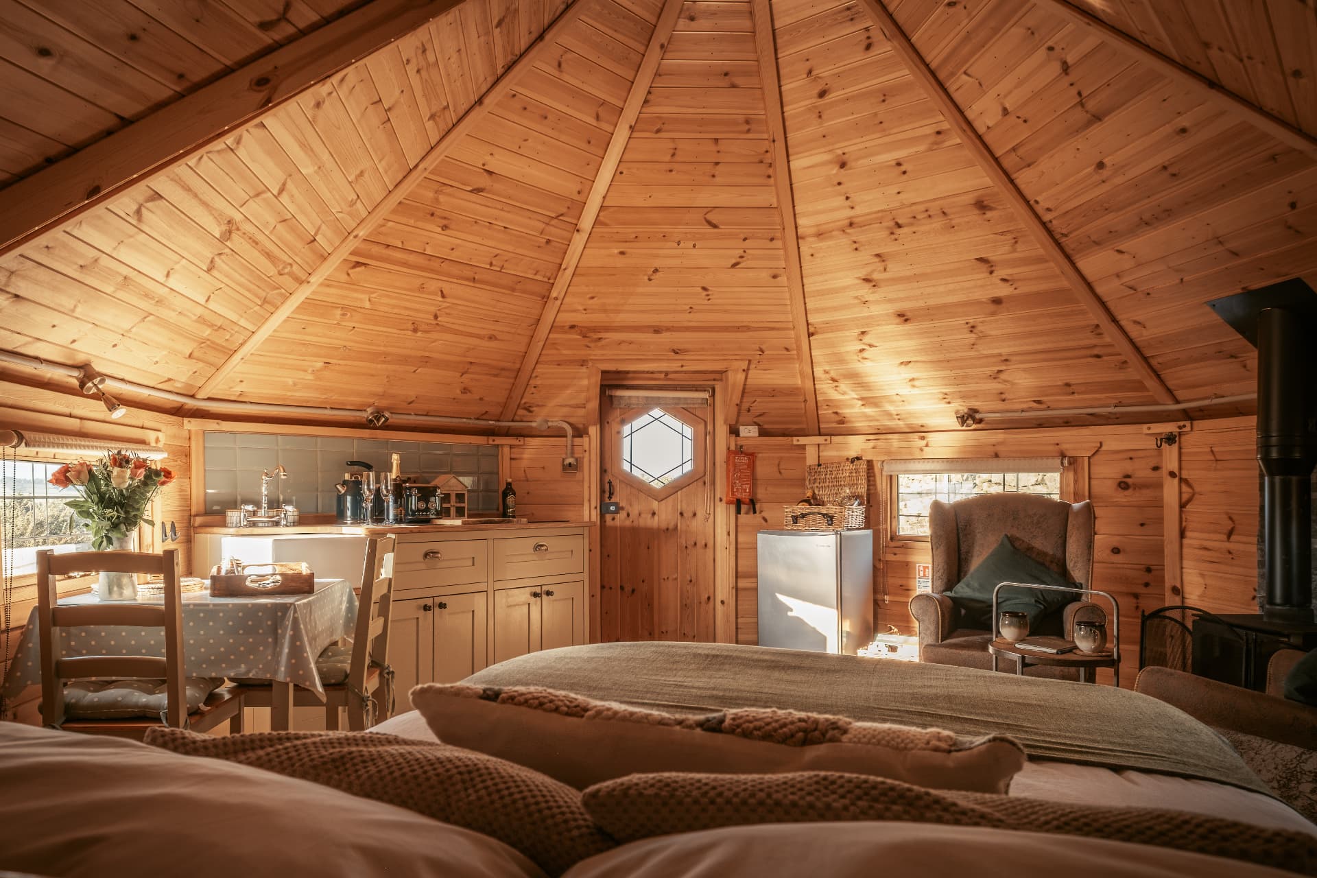 The view from the bed inside The Mallard — kitchen, wingback chair and wood burner beneath the pitched ceiling